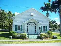 One-story, white, wooden building with front entrance and two windows, both with green shutters