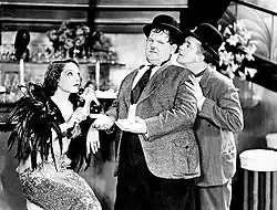 Black and white film still showing Lupe Vélez seated and wearing feathers with Laurel and Hardy in a scene from Hollywood Party (1934)