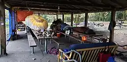 The inside of a wooden picnic shelter in a park with a roof and no walls with people in sleeping bags, couches and various other signs of occupation