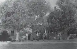 a black-and-white photo of a farmhouse in Utah the 1870s. The house is covered by some trees, and four people stand in front of it
