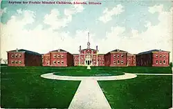 A colored postcard showing a large complex of brick buildings behind a large grassy pavillion