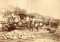 A black-and-white photo of a locomotive carrying and flanked by workers.