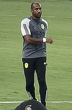 A man wearing a grey Nashville SC training shirt with Adidas track pants stands on a soccer field.