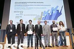 Seven people stand on a stage, with a screen behind them which has projection of a slideshow slide with text and a design on it.