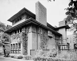 The east and north elevations of the Heller House's facade. There are vines growing on the brick facade. The second floor is topped by a protruding eave, and there is a rectangular brick chimney to the right. Another roof is located above the third story.