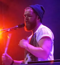 blurry image of Jack Garratt wearing a white print t-shirt and dark blue tuque perched on the back of his head, singing into a microphone onstage