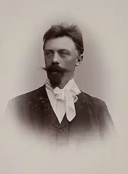 A head and shoulders photo portrait of a forty something man, with a broad pointy moustache and long goatee bread, wearing a white scarf, facing to the left