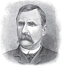 A man with dark hair and a dark mustache wearing a white shirt, black tie, and black jacket