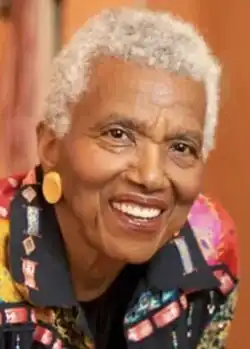 Black woman with grey hair wearing big ear-rings and a multi-coloured jacket