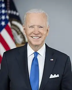 Official presidential portrait of Biden smiling, wearing a navy blue suit jacket with an American flag lapel pin, white shirt, and blue necktie.
