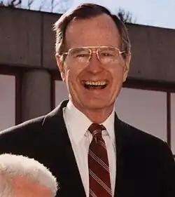 George H.W. Bush, outside in a black suit and eyeglasses, smiling