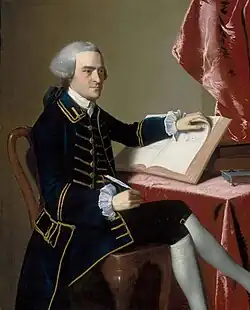 Full-length portrait of a young man seated at a table - he wears a finely tailored dark suit, knee breeches with white stockings, and a wig in the style of a European gentleman. He holds a quill pen in his right hand, and is turning the pages of a large book with the other hand.