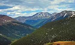 A view of mountainous terrain with a forested valley at the bottom rising to bare slopes with snowfields higher up. In the center one peak stands higher than the others.