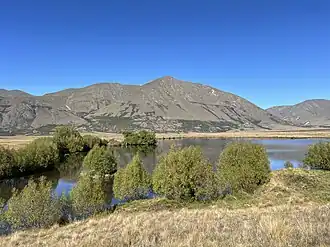 A still lake surrounded by trees and grassy plains, with mountains in the background