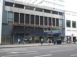 A grey building with blue panels and a dark blue, rectangular sign reading "LANCASTER GATE STATION" in white letters and people in front