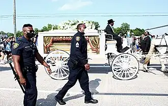 On a street in daytime in fair weather, a white hearse with lustrous yellow curtains with red fringe, topped by a bouquet of white flowers, drawn by white draft horse. A driver in a black suit and black top hat is in the box seat of the hearse. In the foreground, alongside the hearse, are two Houston police officers in duty uniforms. Onlookers are gathered alongside the hearse in the background and walking behind the hearse.