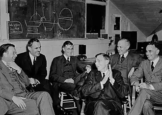 A group of in suits men sit around, laughing. On the blackboard behind is a discarded plan for assembling a uranium core.
