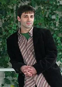 Luis Prieto wearing a light-coloured striped shirt and dark jacket, leaning on right elbow and smiling directly at camera