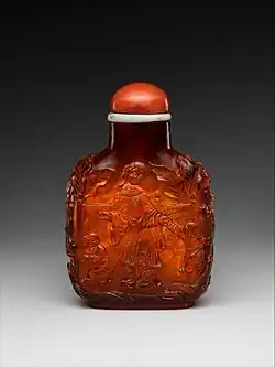 A short amber bottle in a museum display. It features a small coral stopper and a carved motif of a woman and two children walking in a garden