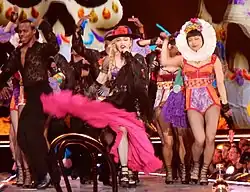 Picture of a group of people dressed in colorful, Mexican-styled clothing. The woman in the middle is blonde, wears a black hat with a pink flower, and waves her pink skirt towards the camera. Her hair is braided and she sings to a microphone in her mouth. To her right, there is a female in an orange and purple leotard, and a white wreath around her head. Also present is a black male in dark clothing.