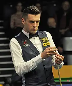 Mark Selby stands looking at a table while he chalks the cue in his left hand.