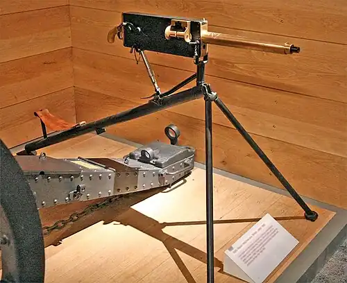 Brass barrelled gun with black coloured rear parts. On a tripod on a wooden plinth with a wood wall behind.