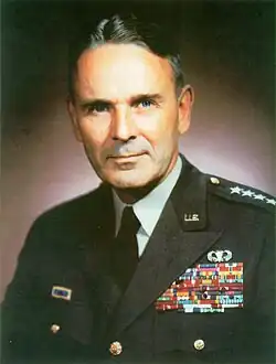 Middle-aged man with graying dark hair parted slightly off center. He wears a green dress uniform, with suit and tie, is clean-shaven, and has four stars on his shoulder to indicate his rank.