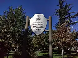 A low-angle, centered shot of a white, shield-shaped sign with Georgian writing and a crest on it. The sign is mounted on two posts and set in a park with green and brown-leafed trees under a clear, deep blue sky.