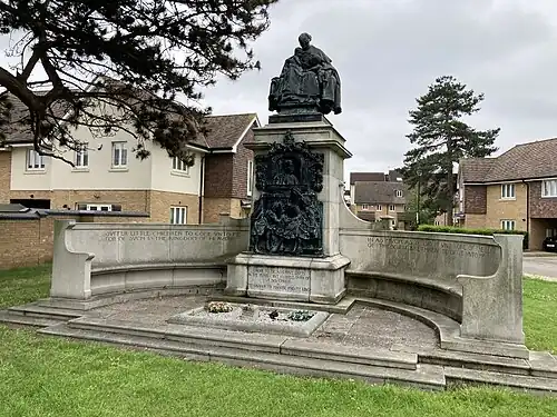 Memorial consisting of two long, curved stone seats attached to a tall stone plinth with a large bronze relief, crowned with a bronze sculpture of a woman with two children.