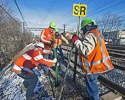 Four men in reflectorized orange vests and neon green helmets standing next to a set of four railroad tracks installing a signpost next to the track. One is holding a sign with the letters "SR" in black on yellow. The ground, covered with a thin snow, slopes steeply to the left and an overhead gantry for the catenary is in the background.