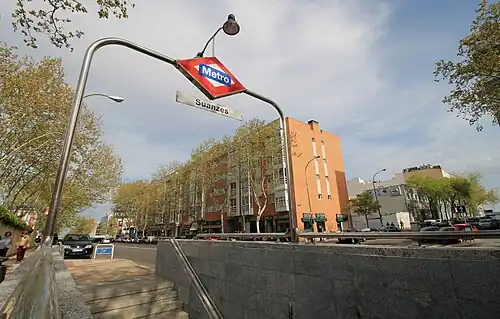 Photograph of a street from the pavement. From a height of about 1 meter we see stairs leading downwards covered by a metallic arch with a rhombus with the Madrid Metro logo and a rectangular sign reading "Suanzes". On the other side of the street, a five-floor building. There are tall trees on boths sides of the street.