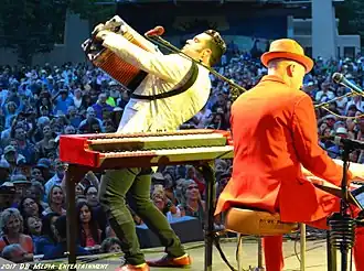 Musician Michael Guerra playing an accordion while facing an audience. To his right is keyboardist Jerry Dale McFadden, playing a piano keyboard while his back is to the camera