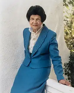 A woman with dark hair wearing a blue suit, leaning on a railing.
