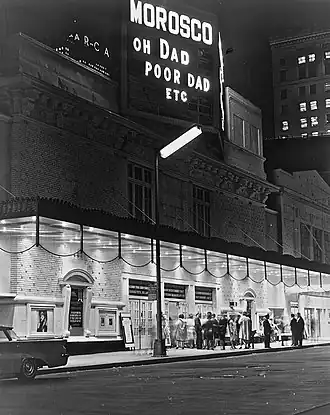 The Morosco Theater as seen from across the street at night. Under the marquee are a group of people. There is a sign displaying the theater's name and the name of the play "Oh Dad, Poor Dad, Etc."