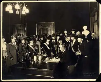 A man sits at a desk signing a document while a large group of women watches