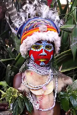 Person in ceremonial outfit with mostly red face paint, a hat with feathers, and shell necklaces