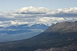A glaciated, flat-topped, gently-sloping mountain shadowed by clouds with a much lower barren mountain in the right foreground.