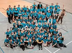 About 100 children of the Musaik orchestra in blue t-shirts are in a gym and are looking up into the camera.