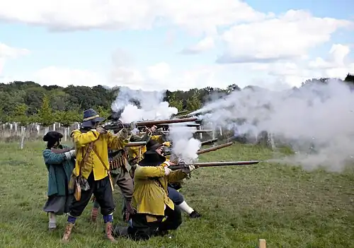 A colour photograph showing a re-enactment of a seventeenth century battle, with a unit of infantry firing muskets