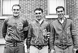 three young men stand side by side. Each has short, dark hair parted in the middle and is wearing an identical team jacket with a stylized maple leaf logo on the left breast.