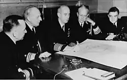 Black and white photograph of five men in naval uniform seated, in front of a table which has on top of it, from left to right, a book and paper, a beeper, an ashtray, a large map and a rotary dial telephone