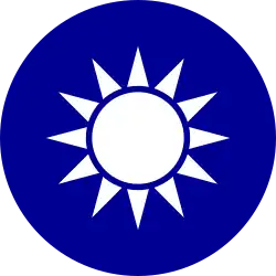 A blue circular emblem on which sits a white sun composed of a circle surrounded by 12 rays.