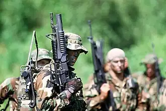 The U.S. Navy SEAL in the foreground is carrying a field radio and is armed with a Colt Model 653 carbine equipped with an M203 grenade launcher.