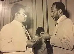 This black-and-white photograph captures a formal interaction between two men in suits. On the left is President Gaafar Nimeiry, wearing glasses and a light-coloured suit paired with a patterned tie, holding a drink in his hand. On the right, Hasan Al-Turabi, in a slightly darker suit and striped tie, engages in conversation and holding a glass. Both individuals are facing each other, appearing deep in discussion. The background shows a simple interior with vertical panelling, and a partially obscured figure is visible in the centre, adding depth to the scene.