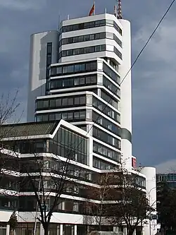A tall modern 15-storey building having a white facade and lots of blue coloured windows with black stripes.