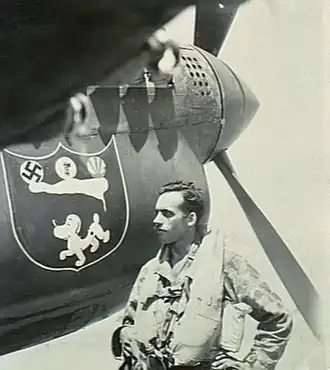Moustachioed man in flying gear standing beneath the nose of a single-engined fighter plane, which pictures a dog and a swastika, among other items