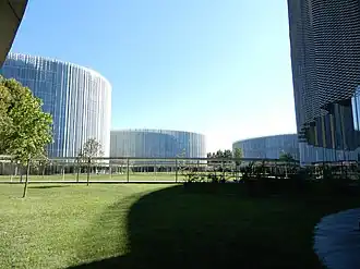 View of the new SDA Bocconi campus in Milan. On the foreground, a green area can be seen. In the background, two white-cladded, circular-shaped buildings can be seen.