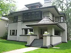Recent exterior view from the northeast corner. In the foreground is the front porch with broad steps and planting piers which hides the front door in shadow. The rest of the house rises behind. A flat yard of turf grass extends up to the base of the house with no other landscaping.