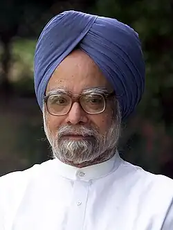 A portrait photograph of a bespectacled Indian man with a dark grey beard, a blue turban, and a white button-down suit with blurred background.