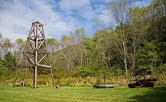 A wooden derrick at left on grass, with trees and a mountain ridge in the background
